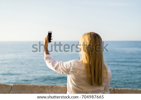 Pretty blond girl using mobile phone at the sea side. Young beautiful woman taking picture on vacations
