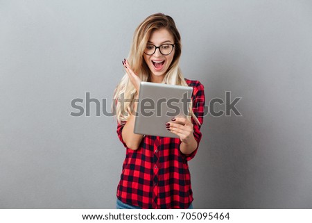 Picture of surprised young blonde woman wearing glasses standing isolated over grey wall. Looking aside chatting by tablet computer.