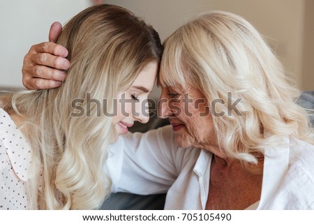 Picture of smiling young lady sitting at home with her grandmother hugging. Looking aside.