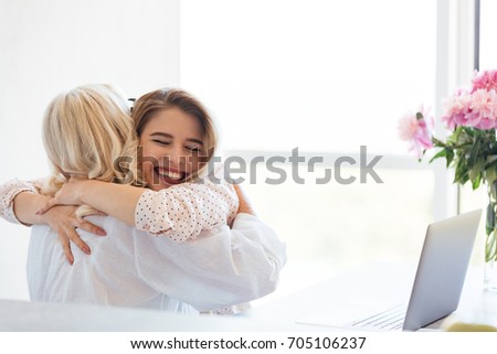 Picture of cheerful young lady sitting at home with her grandmother using laptop computer. Hugging with eyes closed.