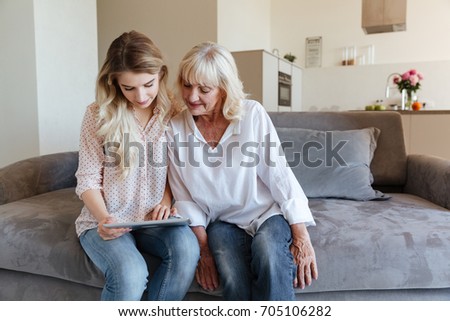 Picture of cheerful young lady sitting at home with her grandmother using tablet computer. Looking aside.