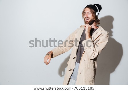 Picture of a handsome hipster man looking aside isolated over wall background listening music with headphones.
