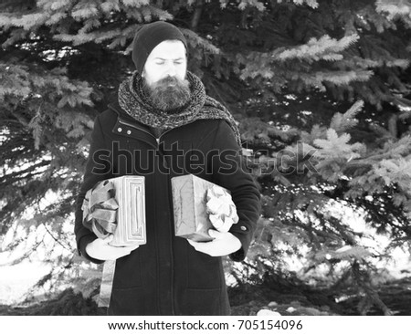 serious bearded man, handsome hipster, with beard and moustache in black hat, coat and scarf smiles with gift boxes as presents on sunny winter day outdoors on natural background