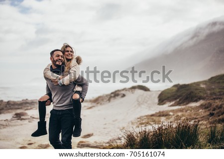 Man carrying girlfriend on his back at winter beach. Young couple enjoying holidays at the seashore.