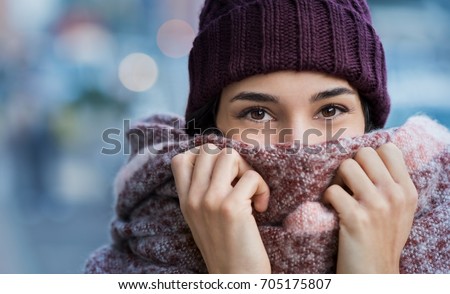 Winter portrait of young beautiful woman covering face with woolen scarf. Closeup of happy girl feeling cold outdoor in the city. Young woman holding scarf and looking at camera. Royalty-Free Stock Photo #705175807
