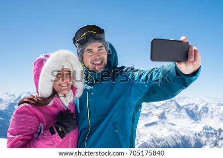 Happy couple taking selfie by smartphone over winter background. Man taking selfie with cheerful girfriend over snowy mountains. Beautiful couple enjoy winter holiday together.