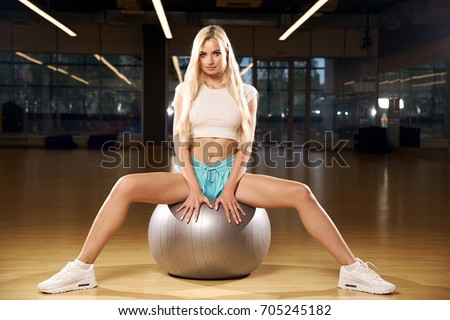 Pretty slim blonde woman with long hair, dressed in white crop top, blue shorts and trainers, sitting on silver exercise ball spreading legs wide apart against two bright lights and gym on background.