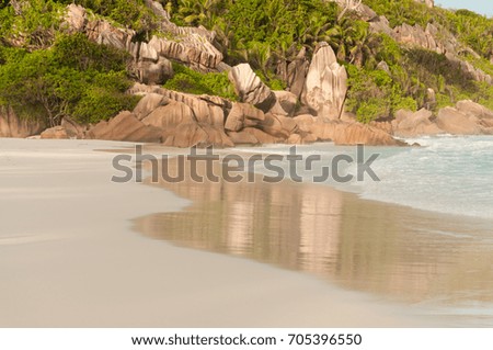 View of the idyllic Anse Coco white sand Beach in La Digue Island, Seychelles, where giant rocks and green vegetation are reflected on the wet sand near the turquoise water.