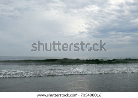 Warm sunlight emerges from the clouds on a cloudy summer day at the beach. The dark shadow of a swelling wave slides across picture plane.