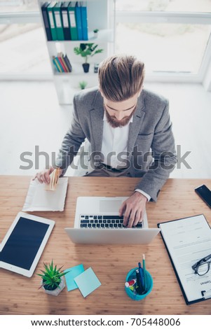 Vertical photo of busy young worker trying to search some information in the internet while having a snack at light modern work place
