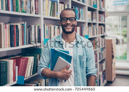 Young cheerful attractive successful african nerdy student is standing with books in the school library archive room, many tomes of ancient textbooks on shelves on the background behind