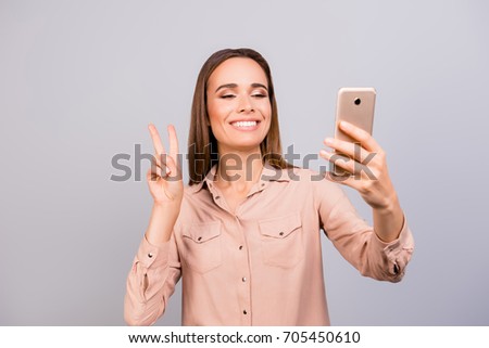 Selfie time! Young brunette woman blogger is making photo for her social networks page, she is posing in a casual smart beige shirt and gesturing peace sign