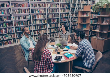International group of four focused clever young students bookworms studying in the ancient university library, sit at the table with books and devices, talk, discuss the project, in casual outfits