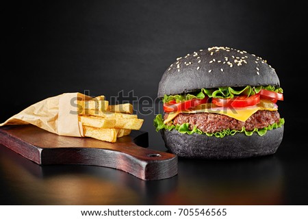 Black burger with french fries on wooden cutting board isolated on black background. Front view.