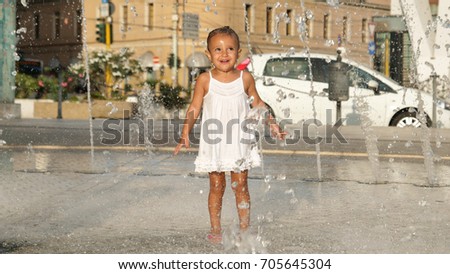 Beautiful little girl having fun and playing in fountain, smiling in white dress, background of transparent water.
