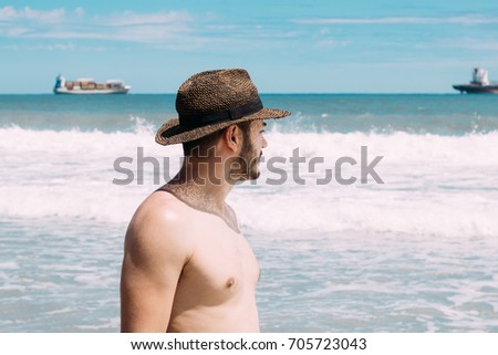 A young man on vacation with sun hat walking by the sea and looking the ships