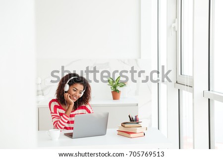 Smiling concentrated african woman using laptop computer by the table near the window