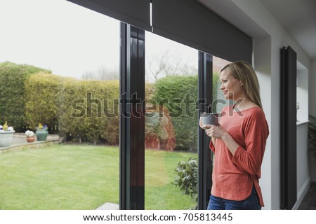 Woman is looking out of her patio door windows with a cup of tea in her hands. 