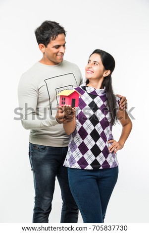 stock photo of Indian smart and cheerful / happy couple holding 3D paper house model and keys, standing isolated over white background, asian couple and real estate concept
