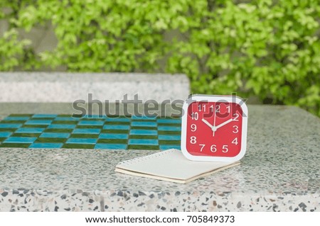 Red alarm clock with note book on marble table in garden.