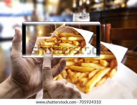 Two hands holding a mobile Smartphone and take a picture of Potatoes fries in a little white paper bag