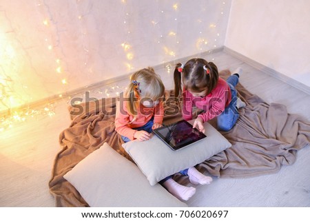 Cute little girls play in computer games on device or watch cartoons, press something on touch screen of tablet, smile and communicate with each other, sitting on rug in bright room with flickering