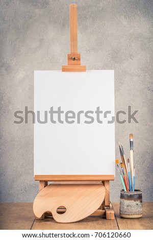 Portable desk easel for painting with canvas blank, brushes and wooden artist's palette front concrete wall background. Retro style filtered photo