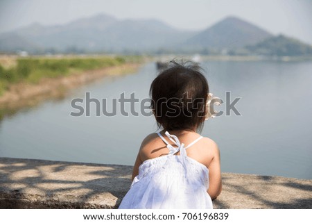 Cute little girl wearing white dress with white plumeria on her ear watching and thinking over reservoir and faraway mountain in the Chonburi, Thailand.