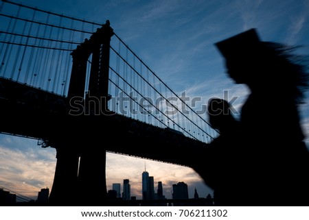 Silhouette of a Woman with long hair in a graduation cap and gown walks passed the iconic Manhattan Bridge in Brooklyn at sunset. Evening sky with silhouette of Manhattan Bridge and the NY skyline.