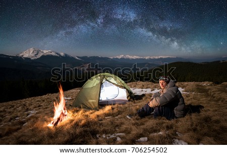 Male tourist have a rest in his camp at night. Man sitting near campfire and tent, looking to the camera under beautiful sky full of stars and milky way. On the background snow-covered mountains