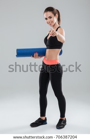Full length picture of happy fitness woman holding fitness mat and pointing at the camera over gray background