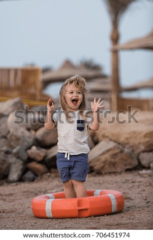 Small kid laughing in orange life ring at beach. Boy child standing with hands up in life buoy. Happiness and expressive emotions. Baby care and childhood concept. Safety on water on summer vacations.