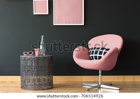 Pink cup, sunglasses and glass bottle on inspiring coffee table next to pink chair against black wall