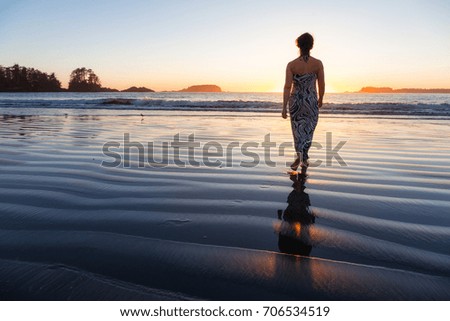 Beautiful woman in a casual dress is walking barefoot on a sandy ocean shore towards the water.  Picture taken on Chesterman Beach, Tofino, Vancouver Island, BC, Canada, during golden summer sunset.