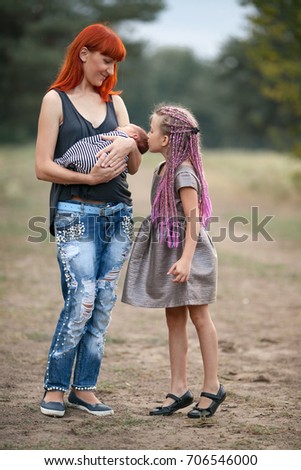 Happy mother with her two children on walk in park. Mother holds in her hands newborn baby, daughter stands next to mam and kisses her little brother.