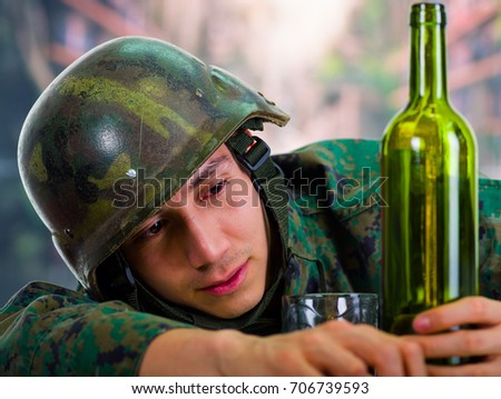 Handsome young soldier wearing uniform suffering from stress post-war, drunk and holding a bottle with one hand, with a glass of ron next to him, in a blurred background