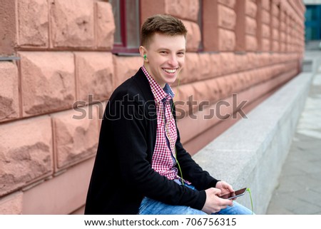 Cheerful happy young man in plaid shirt and jacket listening to music with headphones from cell phone and singing outdoors. Student with smart phone and headphones
