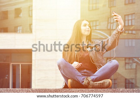 Young brunette girl in jeans doing selfie on a smartphone on the background of an office building. The concept of fashion to shoot yourself on the phone camera