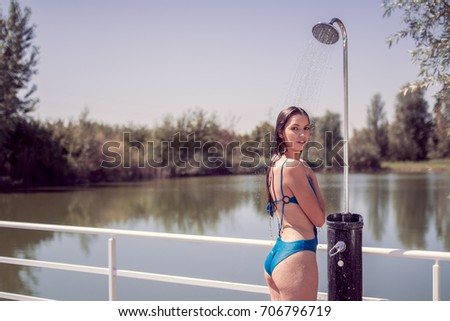 one young adult woman model smiling enjoying, standing, water showering shower, looking to camera, wet long hair, hot sunny day outdoors, lake nature behind, wood floor, one piece swimsuit