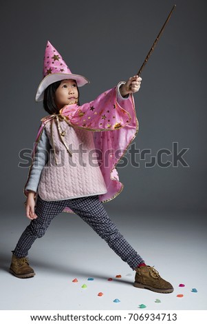 Asian little girl wearing a wizard gown, holding a magic stick, cast a lovely magic. 
 gray background in the studio 