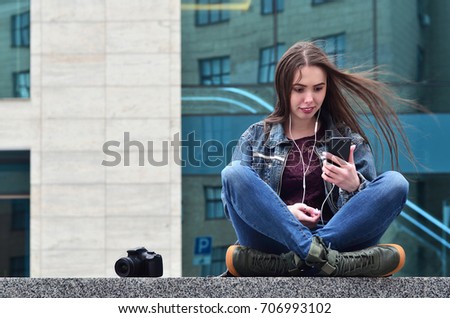A young female photographer in jeans clothes uses a smartphone, sit on a granite parapet against a glazed office building. Concept for lessons or online photography learning and photo-training