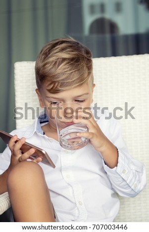Small boy using mobile phone and drinking water. Cute kid watching cartoons on the internet over smart phone and drinking water from glass. 