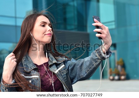 Young brunette girl in jeans doing selfie on a smartphone on the background of an office building. The concept of fashion to shoot yourself on the phone camera