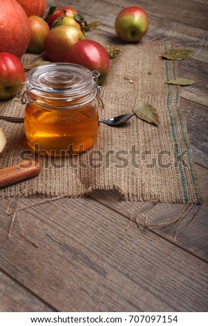 A close-up picture of a colorful composition. Bright autumn fruits next to a glass jar of honey on a wooden table background. A pile of colorful apples next to a spoon and a knife on a rustic napkin.