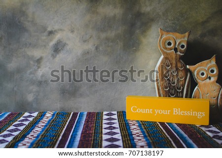 Inspirational yellow wooden sign with words "Count you blessings".  Two wooden owls with Concrete wall in the background.