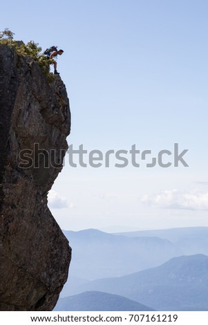 Adventurous male hiker is looking over the edge of a steep cliff on top of the mountain. Picture taken at Lions Peak hike in Lions Bay, North of Vancouver, British Columbia, Canada.