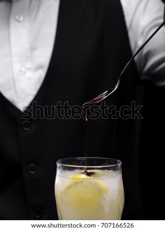 Close-up picture of a professional bartender pouring thick sweet cocktail topping on a fruity alcoholic drink with lemons and crushed ice on the black background. Preparing shake or mix concept.