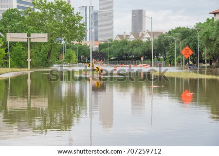 High water rising along Allen Parkway with road warning signs. Residential buildings and downtown Houston in background under storm cloud sky. Heavy rains from tropical storm caused many flooded areas
