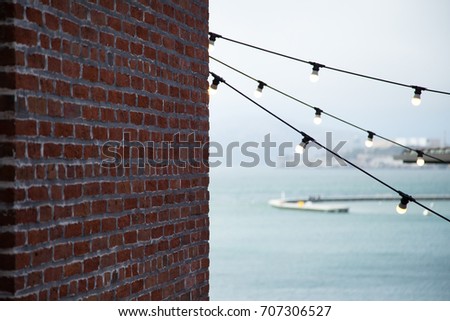 Old brick wall in dark shadow with three strands of light bulbs anchored on corner, with blurry San Francisco harbor in the background