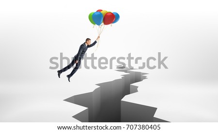 A businessman flies over a huge rift on white surface while holding a bunch of colorful balloons. Crisis management. Business consulting. Avoid trouble. Royalty-Free Stock Photo #707380405